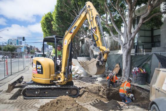 Yellow excavator operates at construction site on city street with workers in safety vests nearby.