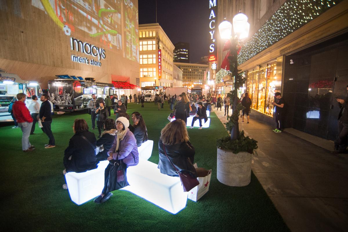Festive holiday street scene with illuminated benches, green turf, and crowds shopping at Macy's in downtown area at night.
