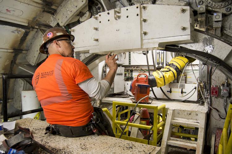 Worker in orange shirt and hard hat performing maintenance on tunnel boring machine equipment in underground shaft.