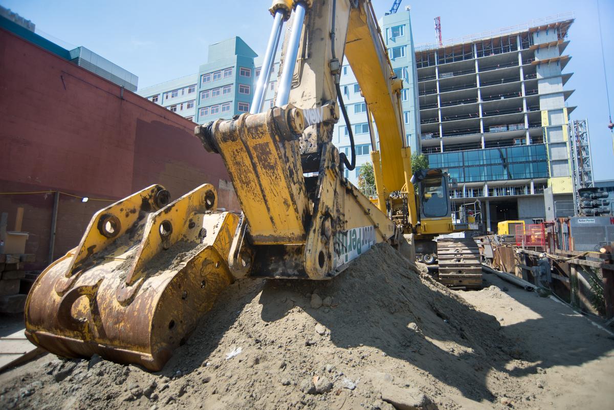 Yellow excavator with bucket attachment on construction site surrounded by apartment buildings and work barriers.