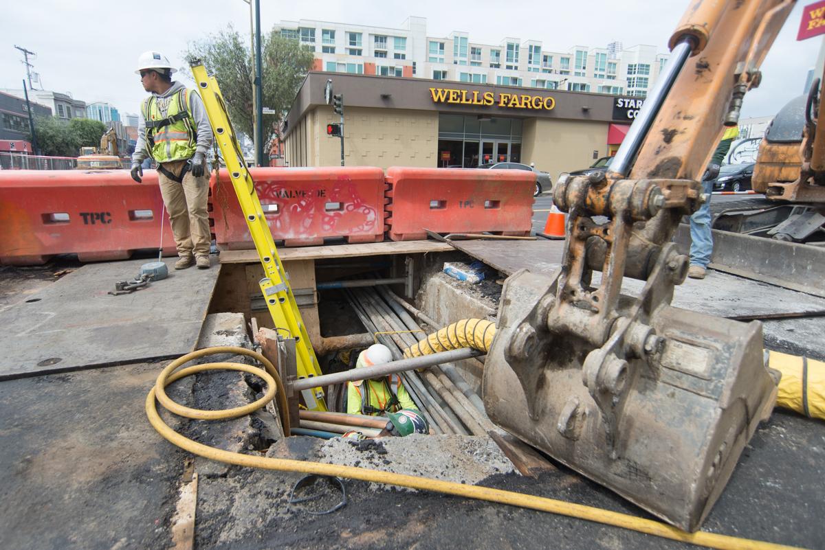 Worker in safety gear at underground utility excavation site near Wells Fargo building during Central Subway construction.