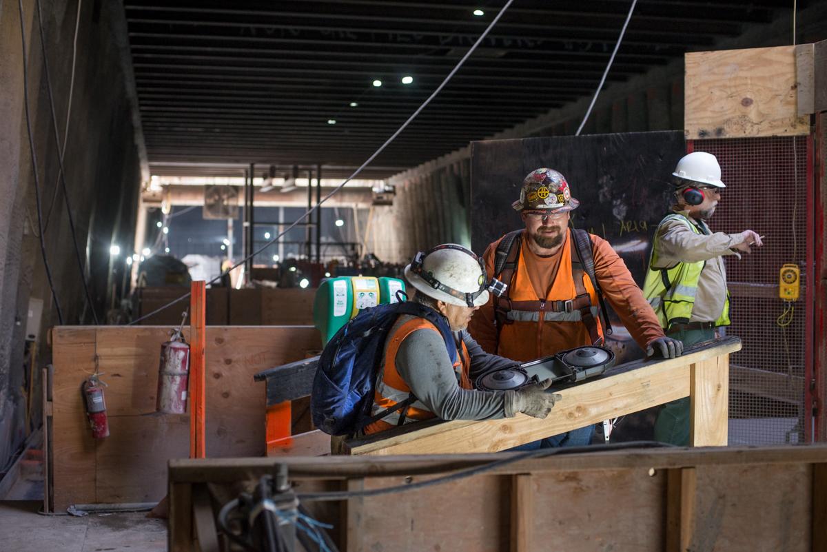 Construction workers in hard hats and safety vests work on structural elements in an underground transit station concourse.