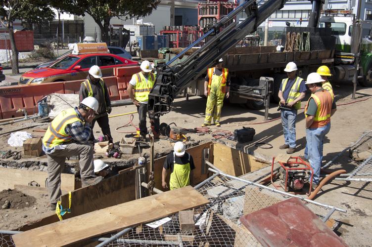 Construction workers in safety vests and hard hats install a water line in an excavated pit on 4th Street, with trucks and equipment nearby.