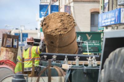 Construction truck discharging excavated soil at transit project site with workers in safety vests nearby.