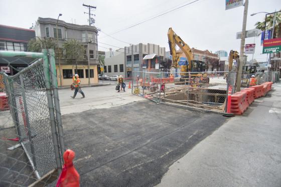 Construction site with fencing, excavator, and red barriers on urban street with buildings in background.