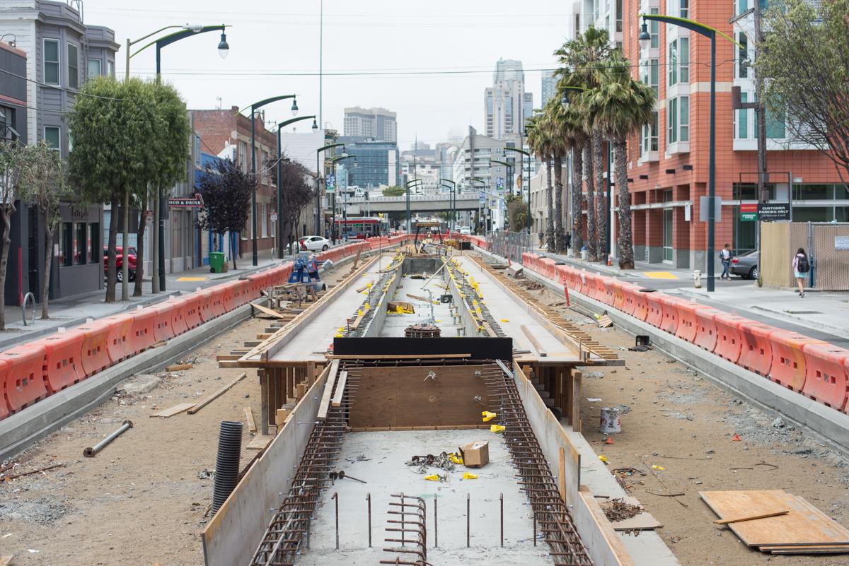 Construction work on transit tracks with concrete forms, reinforcing steel, and red safety barriers lining an urban street.