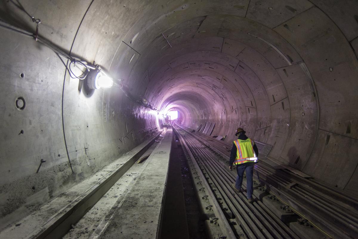 Worker in safety vest inspecting railroad tracks in underground transit tunnel with purple lighting.