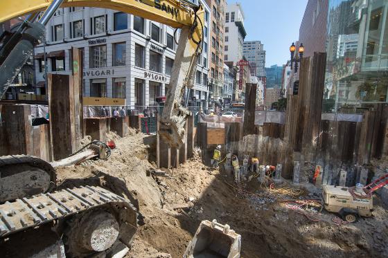 Construction excavation at Stockton and Geary streets with wooden pilings, heavy equipment, and San Francisco buildings in background.