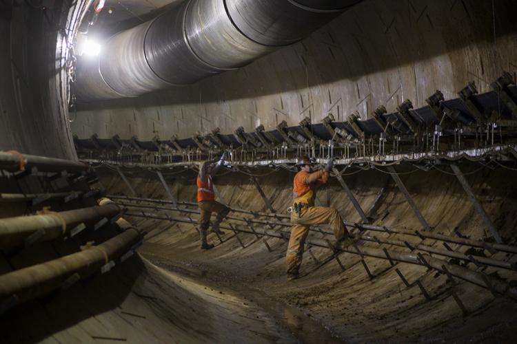 Workers in safety gear operate a conveyor system inside an underground tunnel during transit construction.