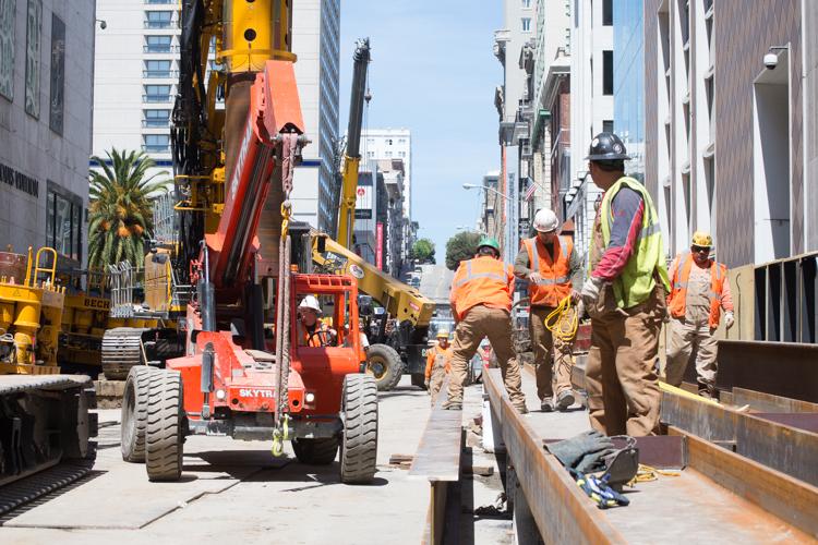 Construction workers and equipment on an urban street with tall buildings, including a red crane and orange machinery.