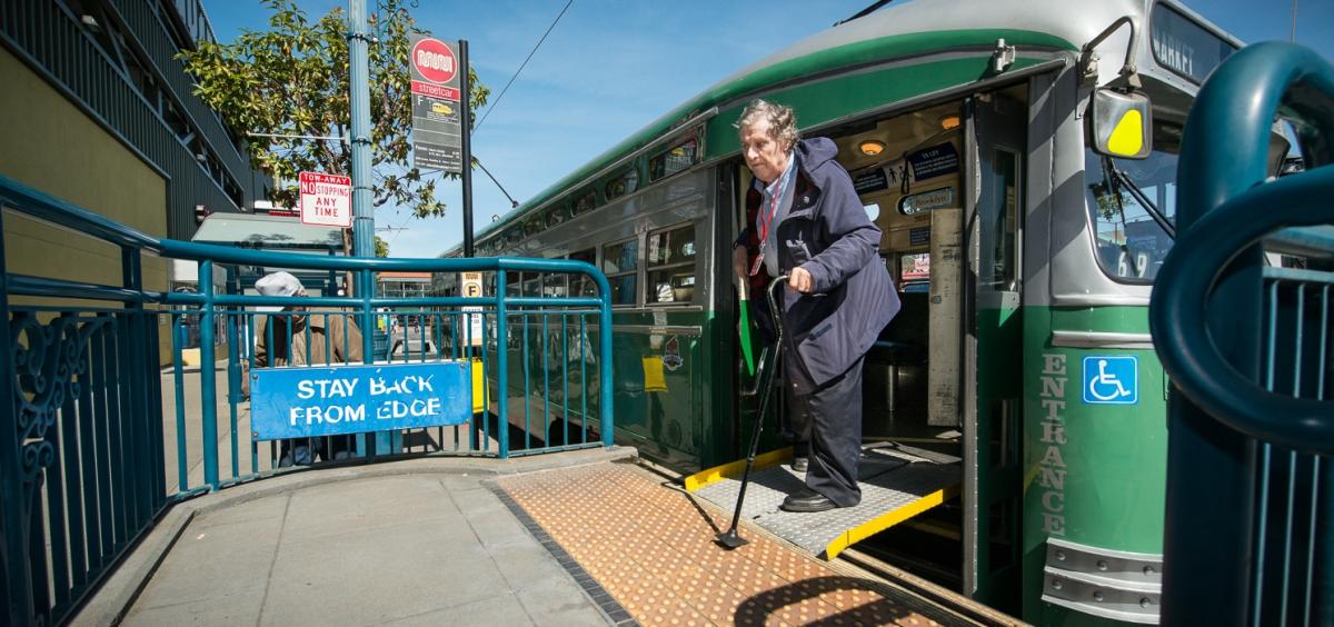 Elderly passenger exiting green and white streetcar using wheelchair ramp at transit stop.