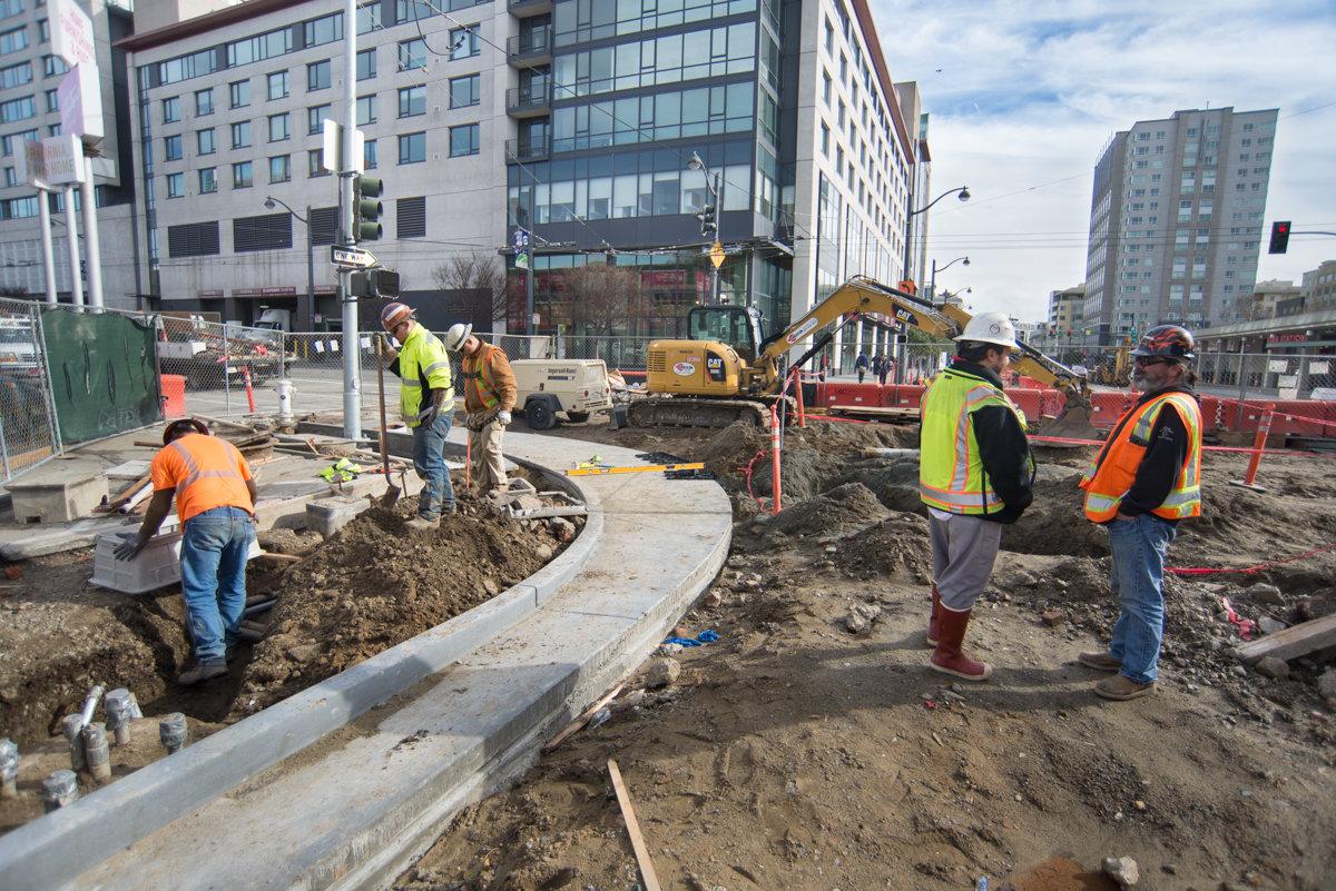 Construction workers in safety vests install curb and gutter on urban street with excavator and buildings in background.