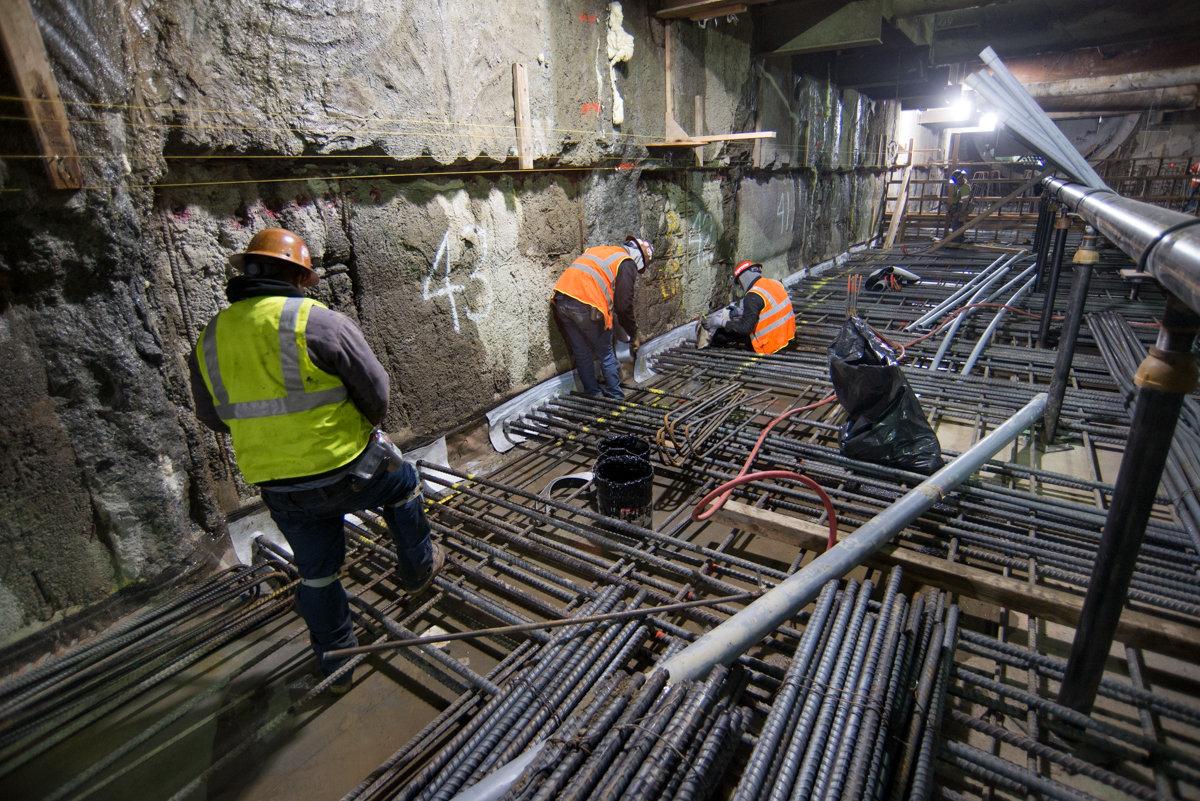 Workers in safety vests install reinforcing steel bars in underground station construction.