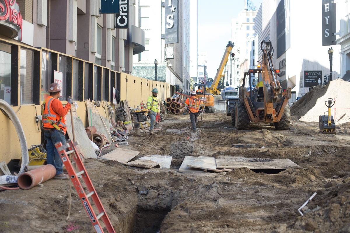 Construction crew excavating utilities along downtown street with heavy equipment and barriers.