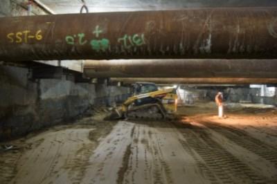 Bulldozer levels soil beneath steel support beams inside underground transit station box.