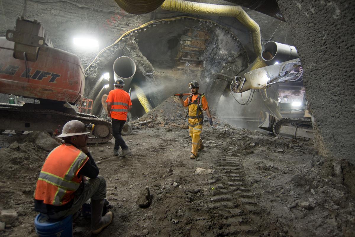 Workers in safety gear excavate underground tunnel with large drilling equipment and dust suppression systems.