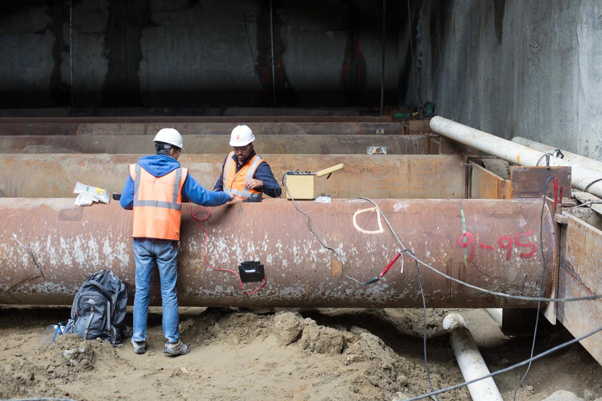 Two workers in hard hats and safety vests inspect a large rusted steel pipe during underground construction work.