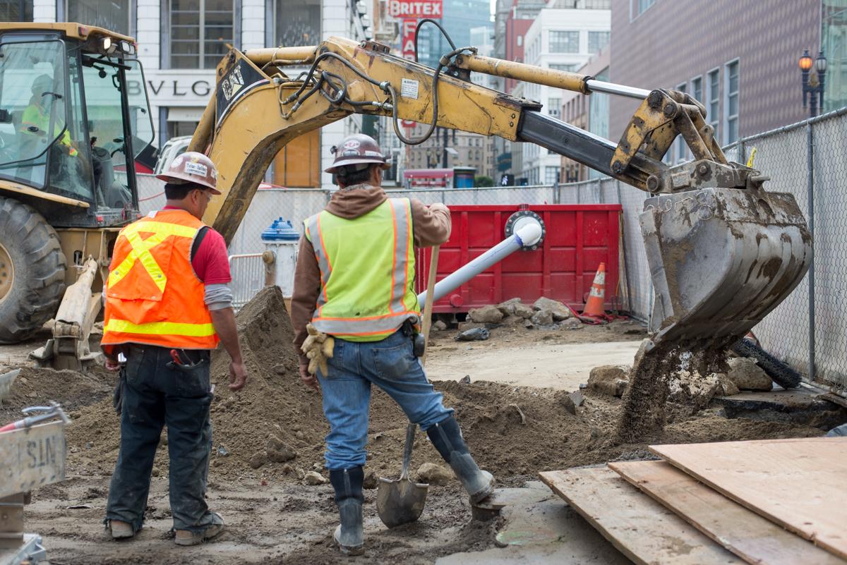 Construction workers operate excavator to backfill trenches at urban transit station site.