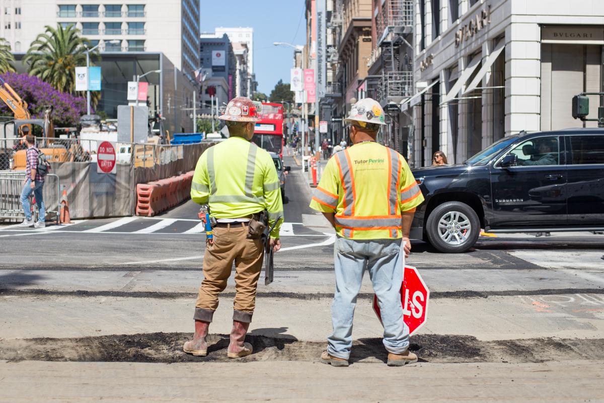 Two construction workers in safety vests and hard hats stand on O'Farrell Street at an active construction site with urban buildings in background.