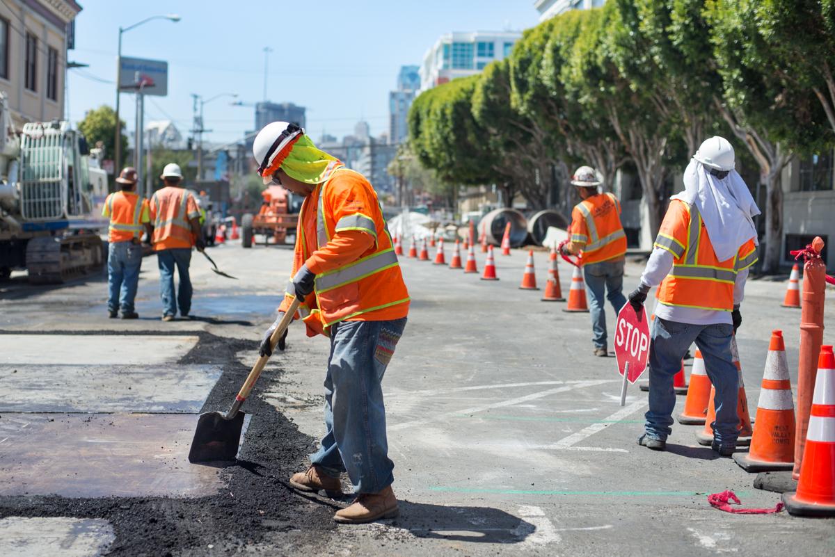 Construction crew performing sewer line installation work on urban street with traffic cones and safety equipment.