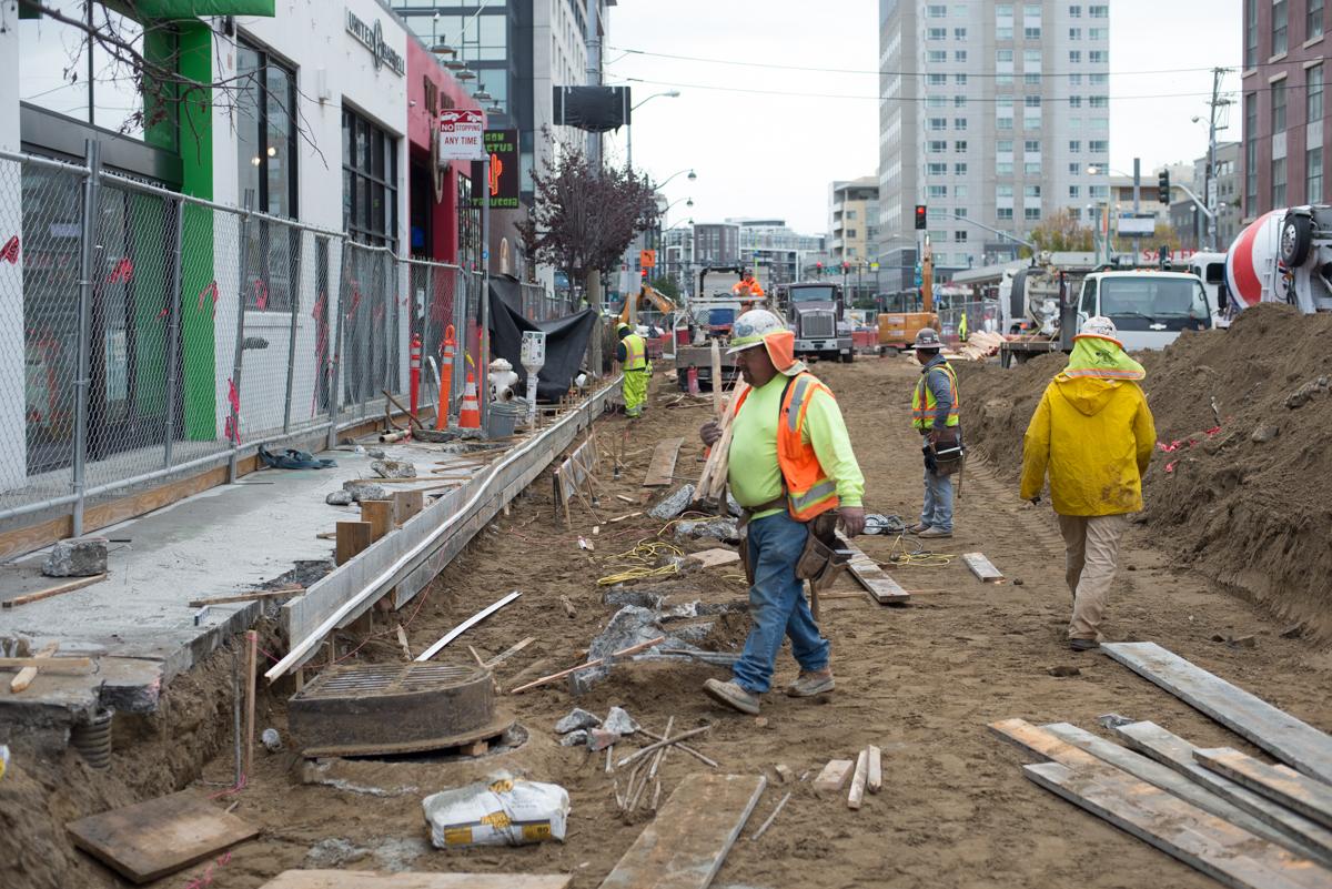 Construction workers in safety vests and hard hats work on an excavated street site in an urban area with buildings and equipment visible.
