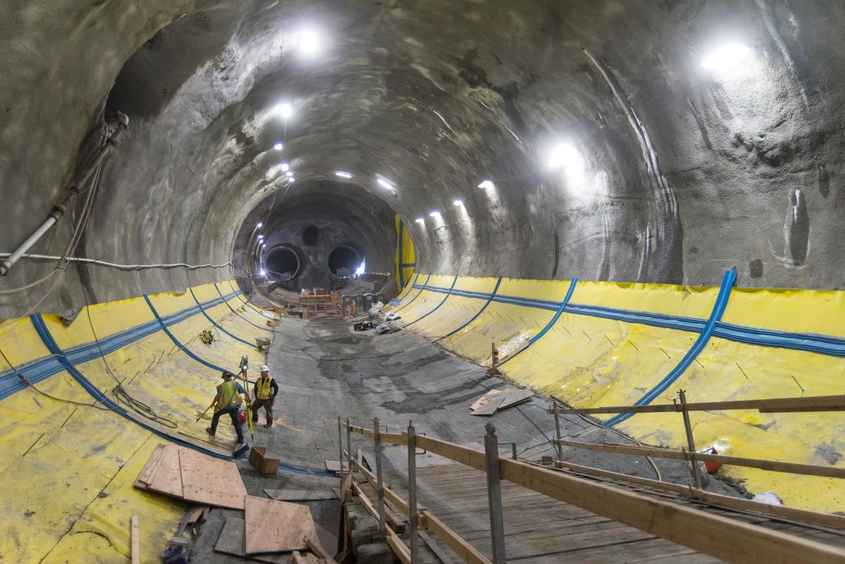 Construction workers inspect underground transit tunnel with yellow safety barriers and blue utility lines installed along concrete floor.