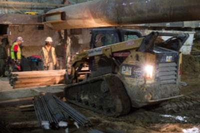Workers load lumber onto a Bobcat skid-steer loader at a transit station construction site.