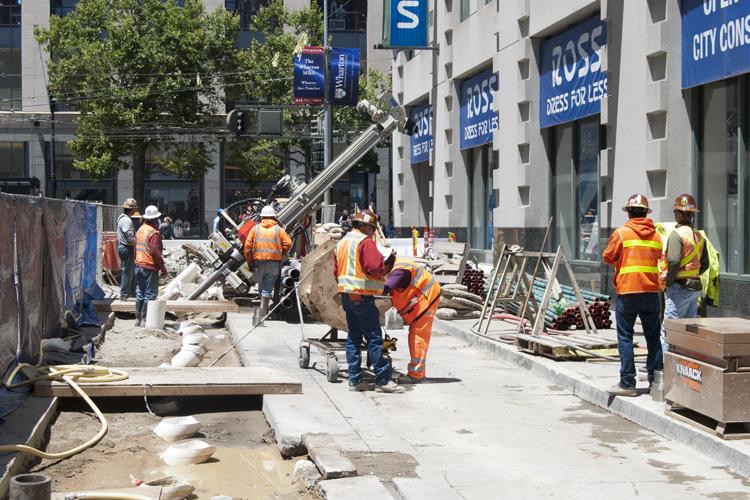 Construction workers in safety vests operating equipment to install subsurface pipes on an urban street.