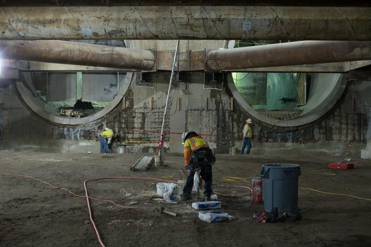 Workers in safety vests perform excavation and preparation work beneath large concrete tunnel structures at a transit station.