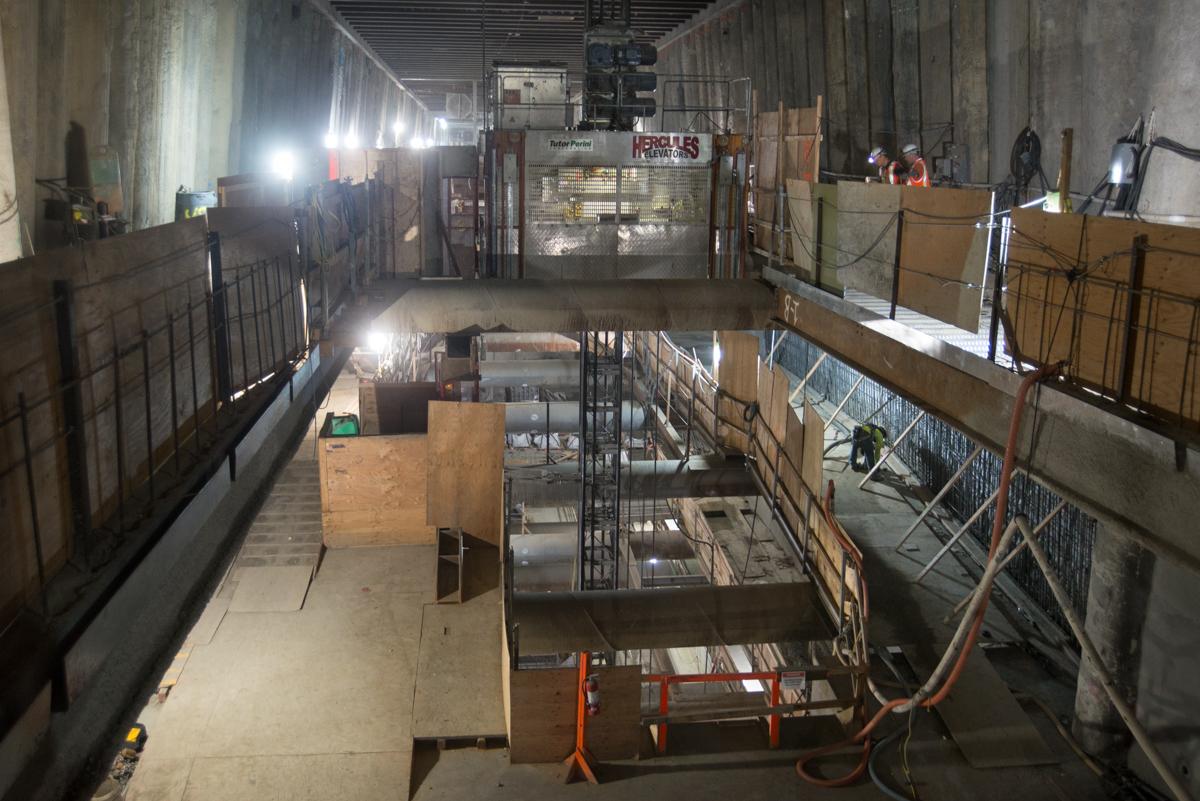 Underground transit station under construction with multiple levels, metal framework, and work lighting visible.