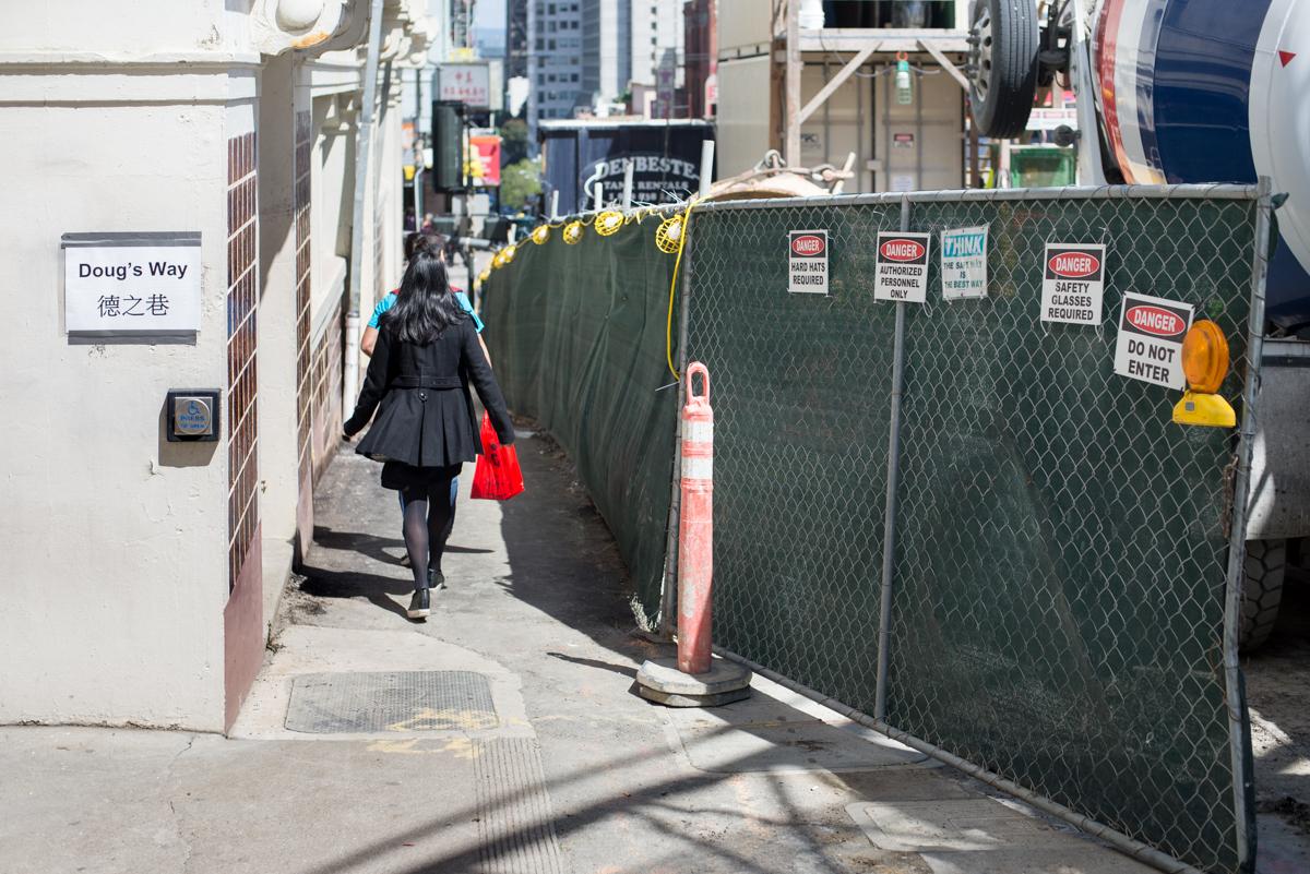 Pedestrian walking through fenced construction area on narrow alley between buildings in urban setting.