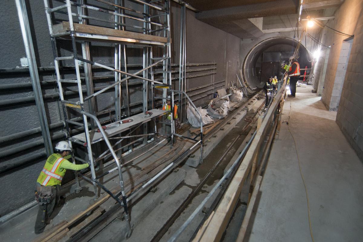 Workers in safety vests install metal studs on underground transit station platform using scaffolding and equipment.