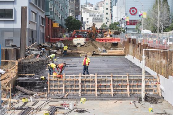 Construction crew installing rebar reinforcement on roof deck at urban street site with safety barriers and equipment.