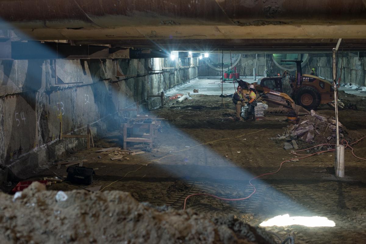 Underground station construction site with excavation equipment, temporary bracing, and light streaming through headhouse portal.