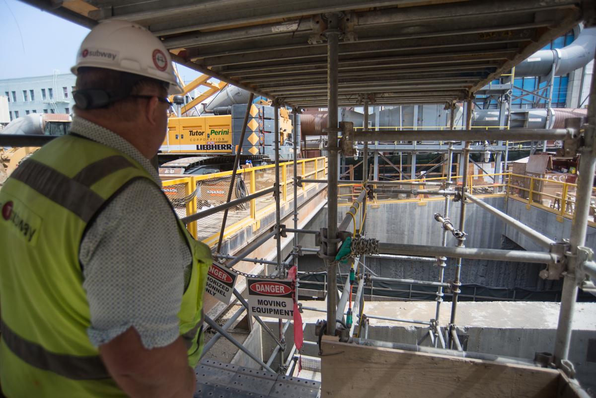 Construction worker in safety gear observing main access shaft excavation site with heavy equipment and scaffolding.