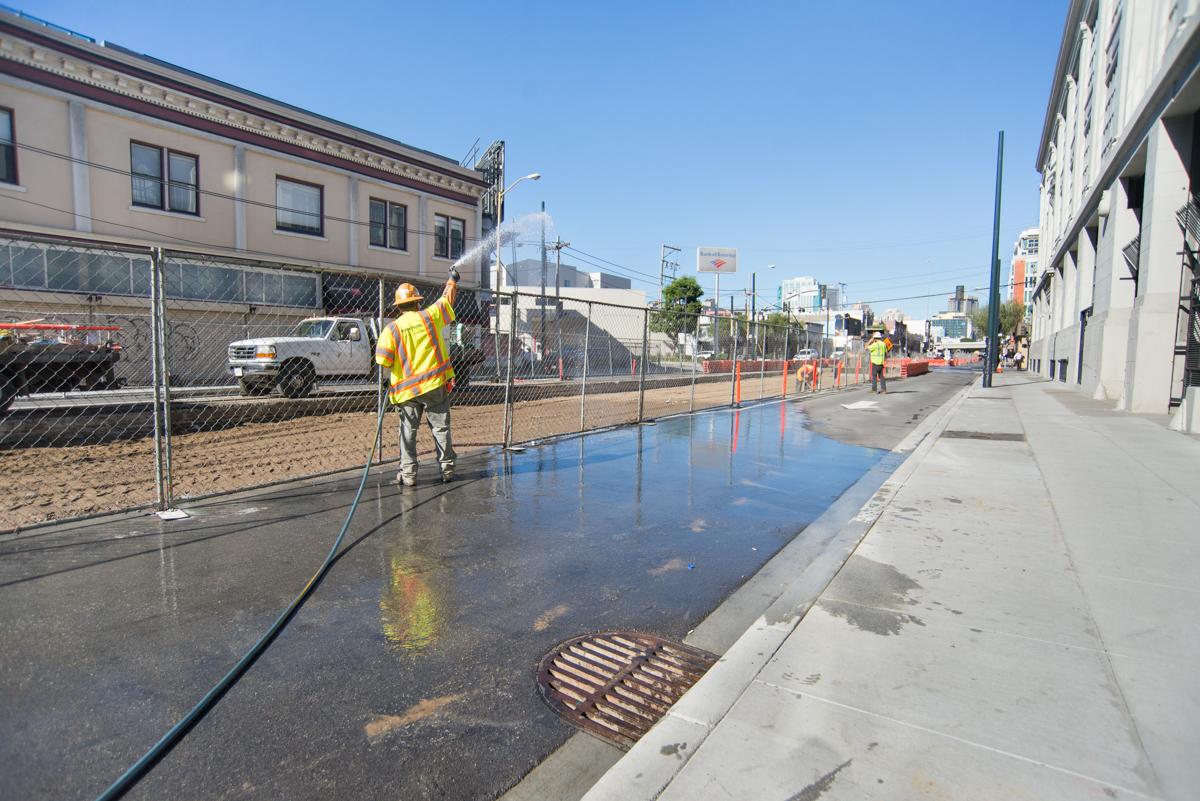 Worker in yellow vest spraying water on street near baseball field to control dust during daytime construction.