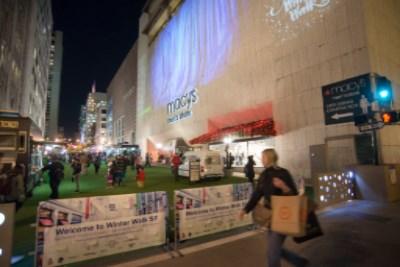 Shoppers and pedestrians gather on a green lawn at night during Winter Walk, with decorated banners, food vendors, and holiday lights visible.