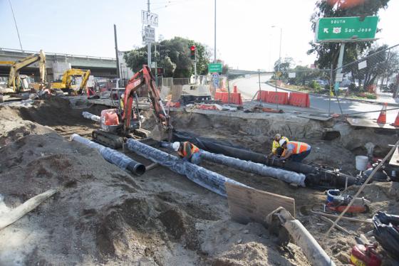 Workers install underground utility pipes at street construction site with heavy equipment and safety barriers.