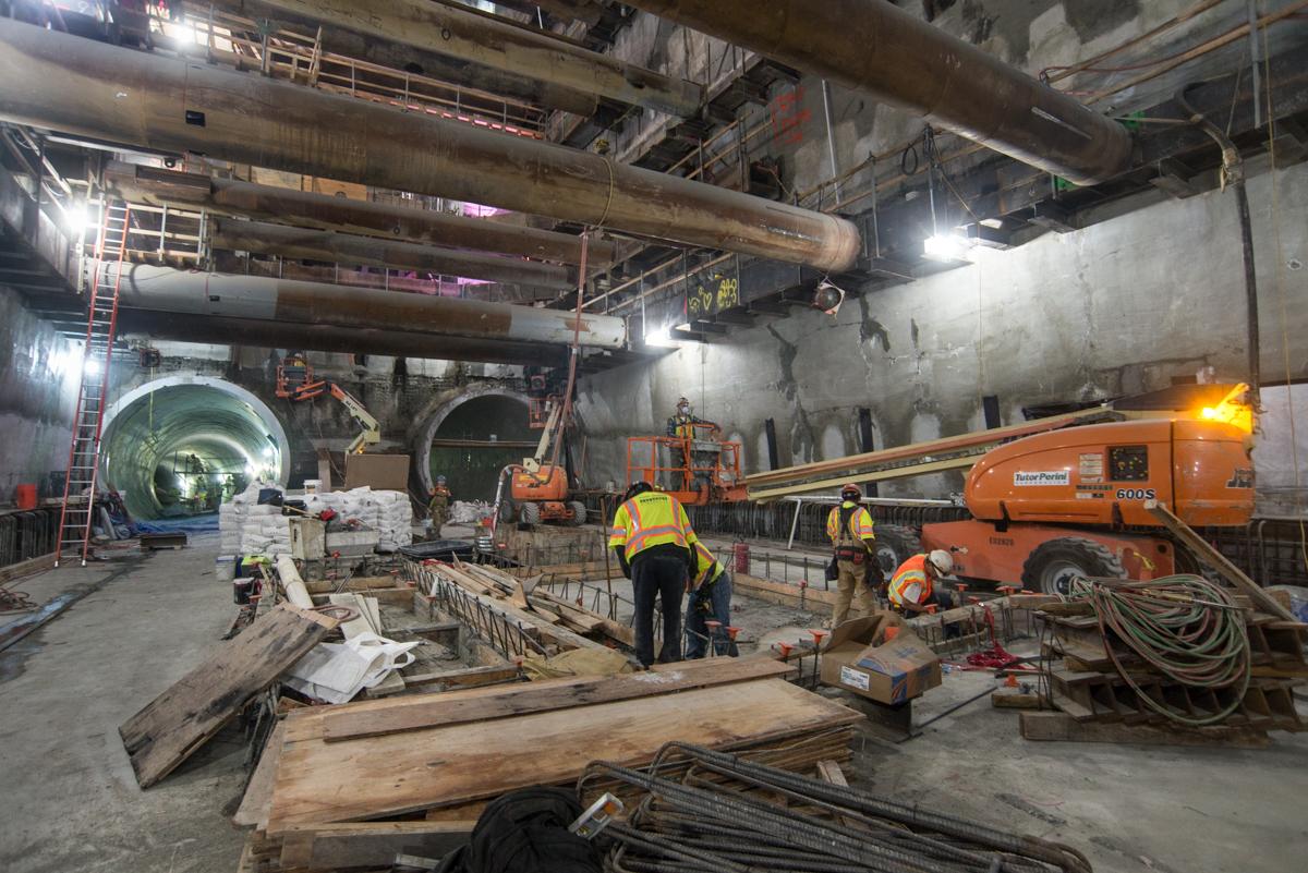 Construction workers in safety gear work on Union Square/Market Street station platform inside a tunnel with large machinery and equipment.