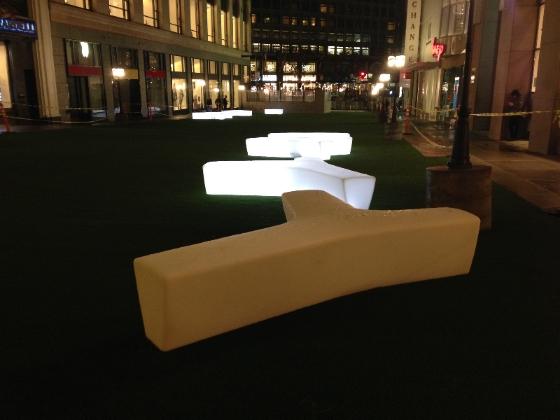 Illuminated curved seating and glowing benches on green turf in urban plaza at night.