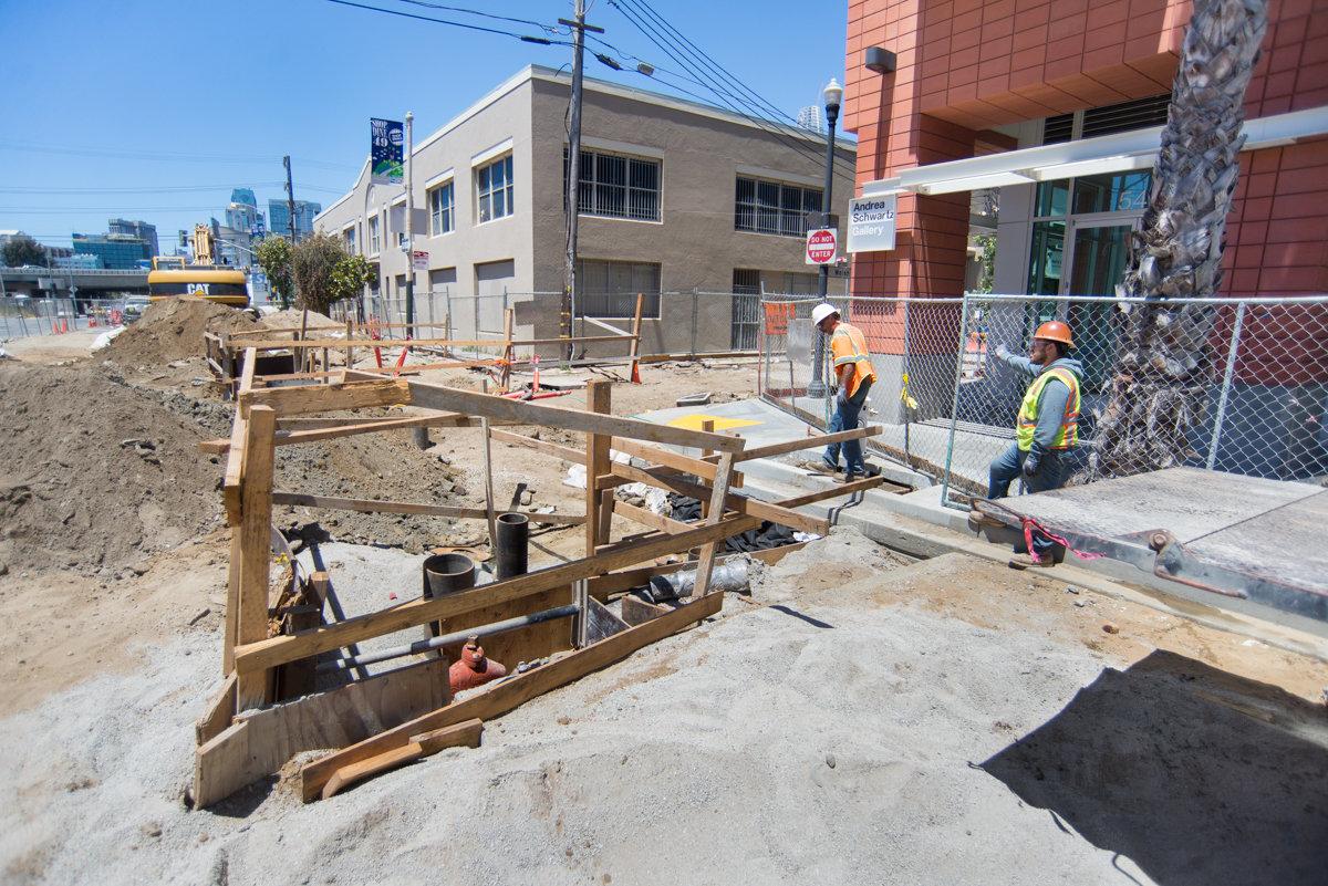 Construction workers in safety vests install utilities along 4th Street near residential and commercial buildings.