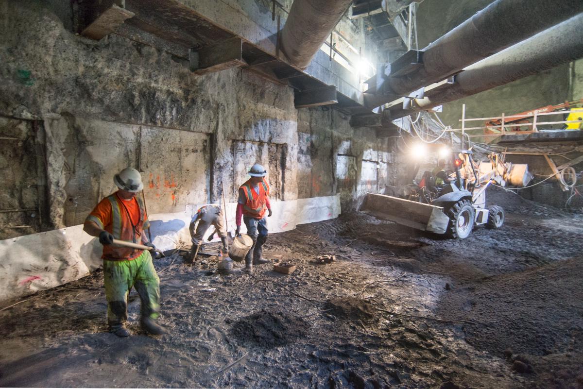 Workers in hard hats and safety vests perform grading work in an underground station cavern with heavy equipment and lighting.