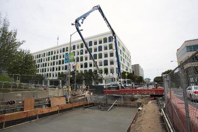 Concrete pump truck pouring roof deck at underground construction site with office buildings in background.
