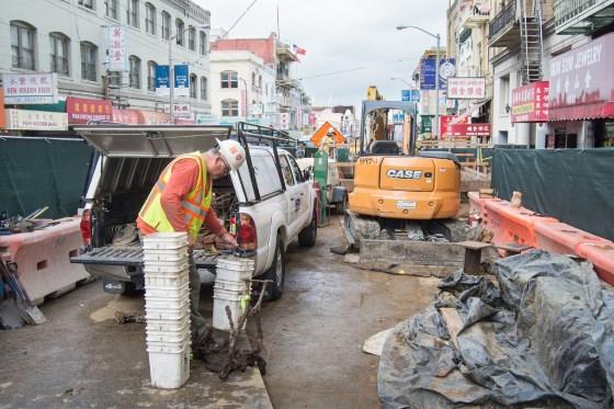 Worker in safety vest unloads pipe from white van during street construction in San Francisco's Chinatown district.