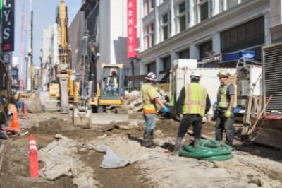 Construction workers perform grouting work on Stockton Street in downtown San Francisco amid urban buildings and equipment.