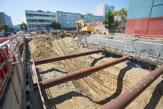 Construction site with large metal pipes and excavation equipment, temporary support structures visible in sandy ground.