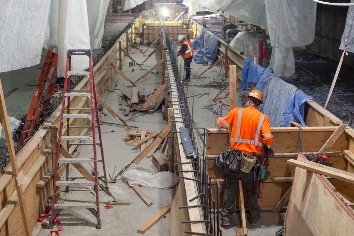 Construction workers in safety gear build wooden forms inside an underground transit station box.