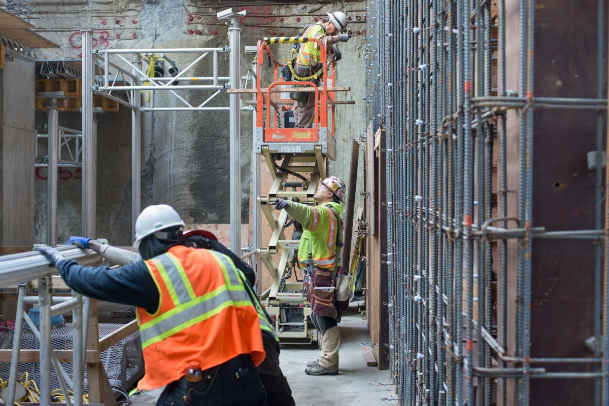Construction workers in safety gear install rebar and concrete forms inside a transit station headhouse using an elevated work platform.