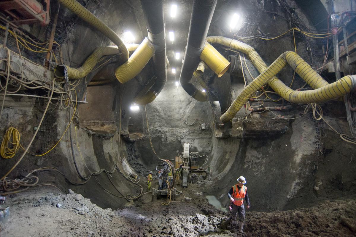 Worker in hard hat and safety vest inside underground tunnel with yellow ventilation ducts and LED lighting during subway construction.
