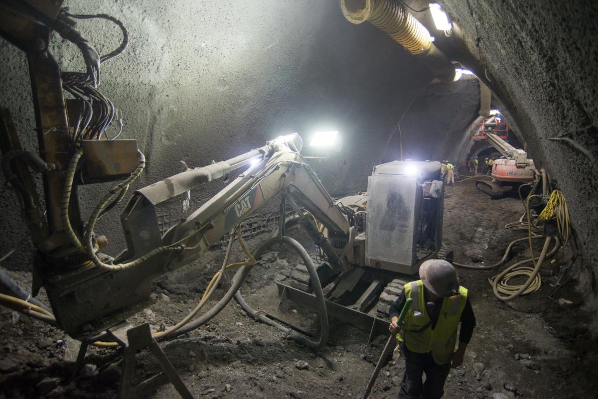 Workers and equipment excavating underground platform cavern with bright work lights illuminating the concrete tunnel walls.